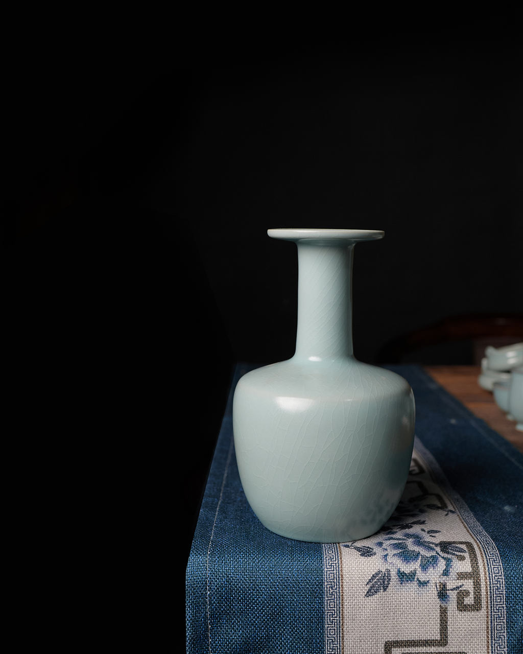 Celadon vase displayed on a blue patterned table runner against a dark interior backdrop.