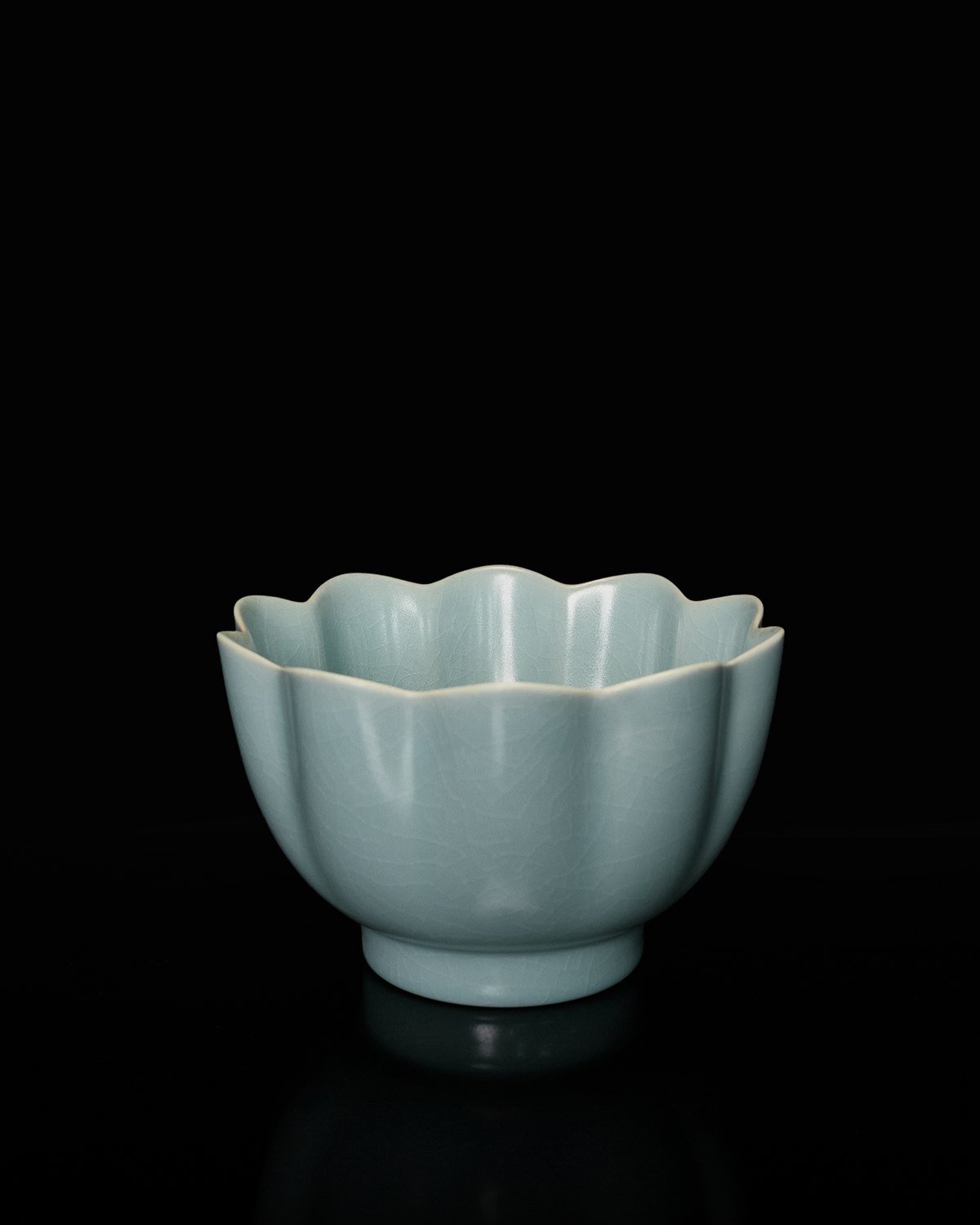 Full silhouette of the celadon lotus bowl on a dark reflective surface with its scalloped rim visible.