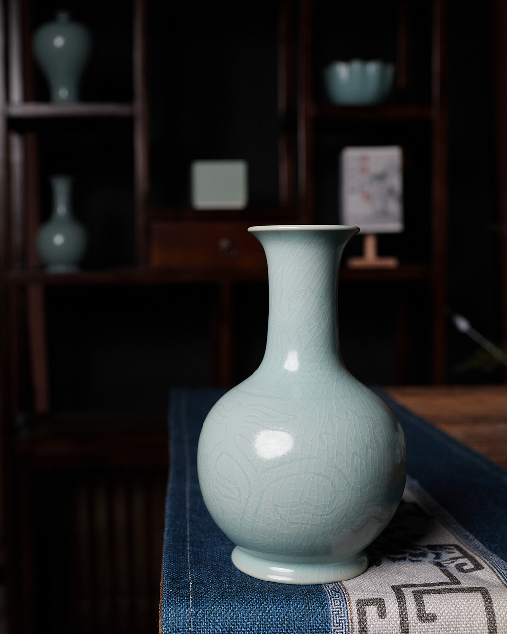 Chinese celadon vase displayed on a patterned textile in a dark traditional-style interior.