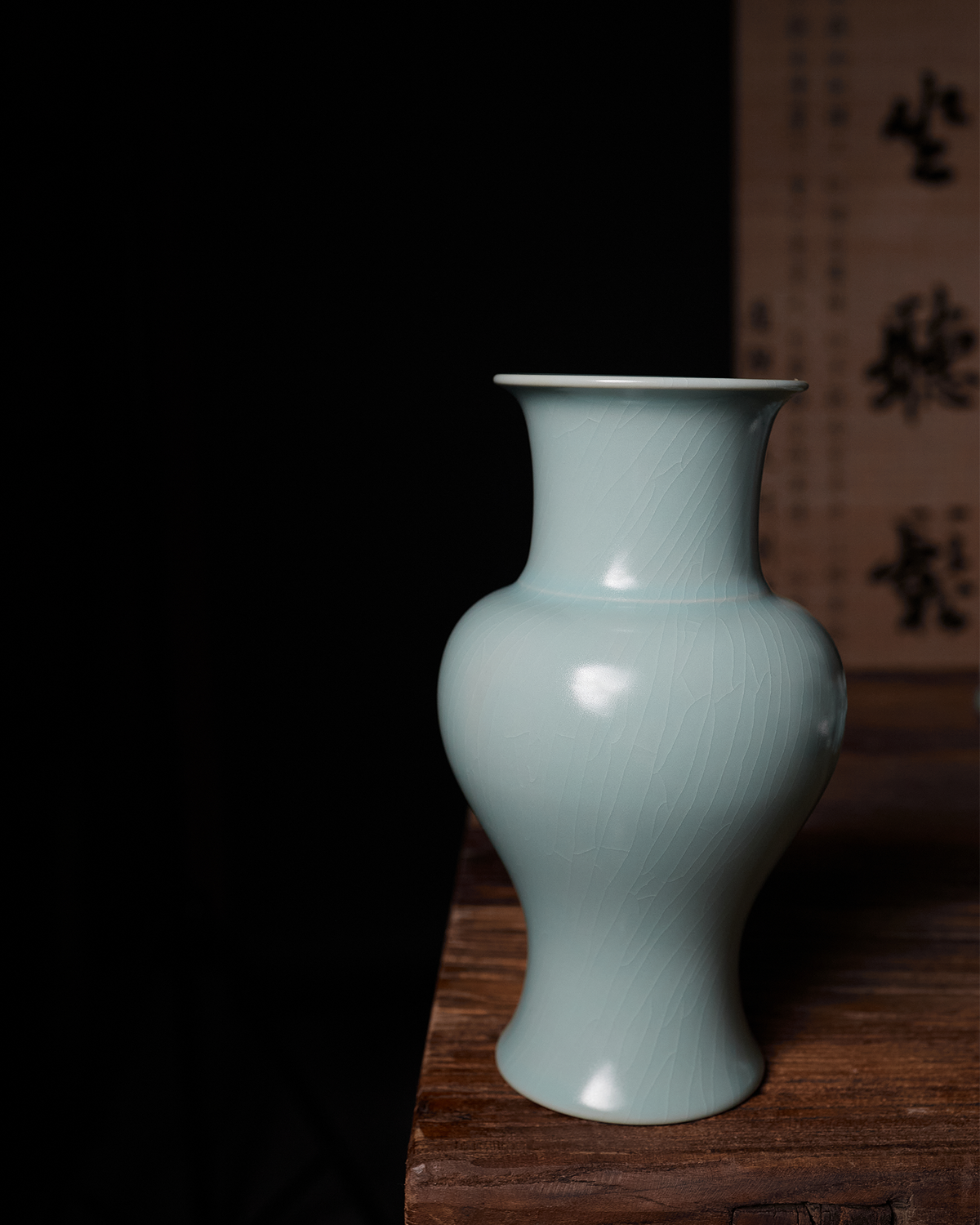 Celadon Guanyin vase displayed on a wooden table beside a softly lit calligraphy backdrop.