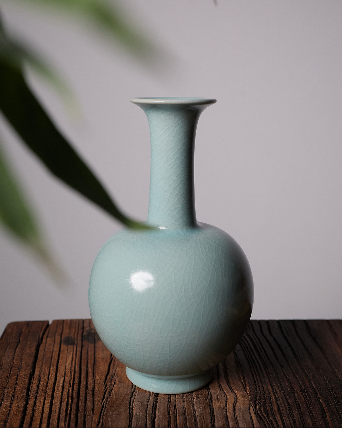 Celadon long neck vase styled on a dark wooden table with a pale wall and soft green foliage.