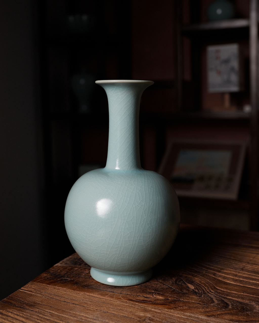 Celadon long neck vase displayed on a wooden table in front of dark shelving.