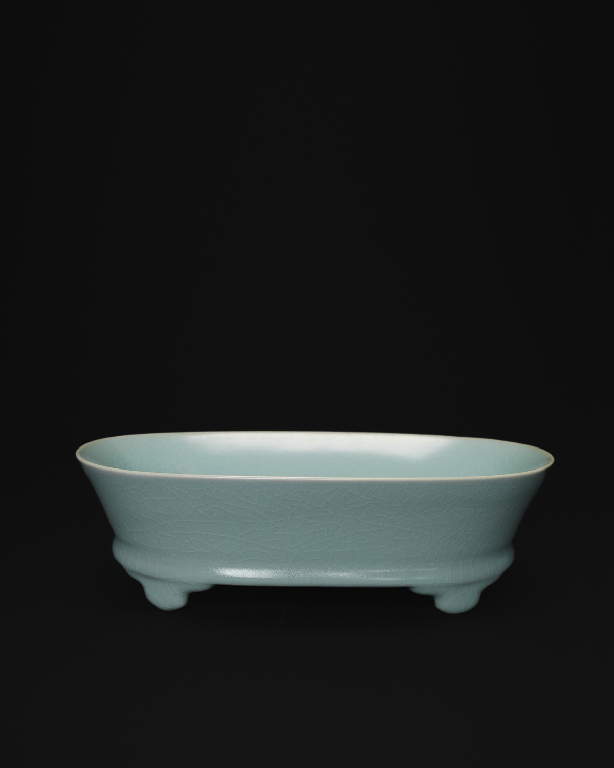 Front view of a celadon narcissus basin with an oval body and raised feet on a black background.