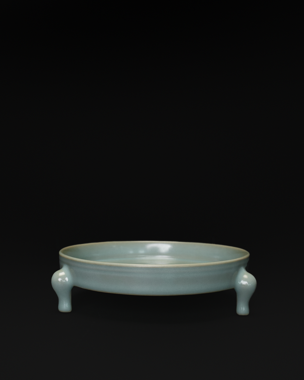 Front view of a celadon tripod bowl with a shallow round basin and three sculpted feet.