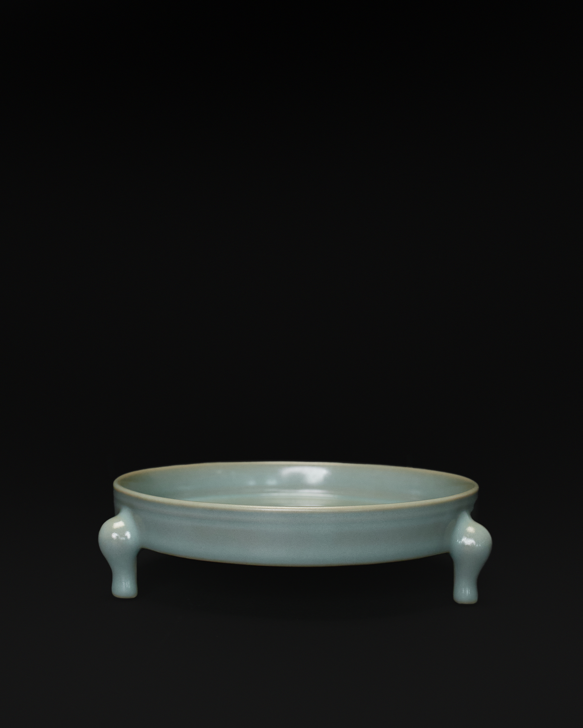 Front view of a celadon tripod bowl with a shallow round basin and three sculpted feet.