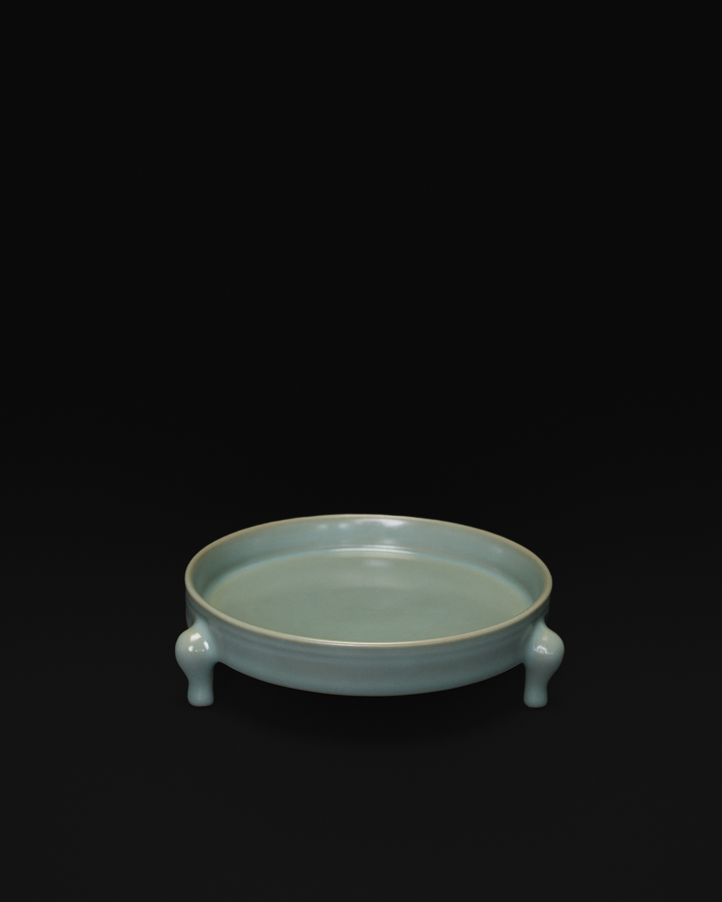 Three-quarter view of the celadon tripod bowl showing its shallow interior and three feet.