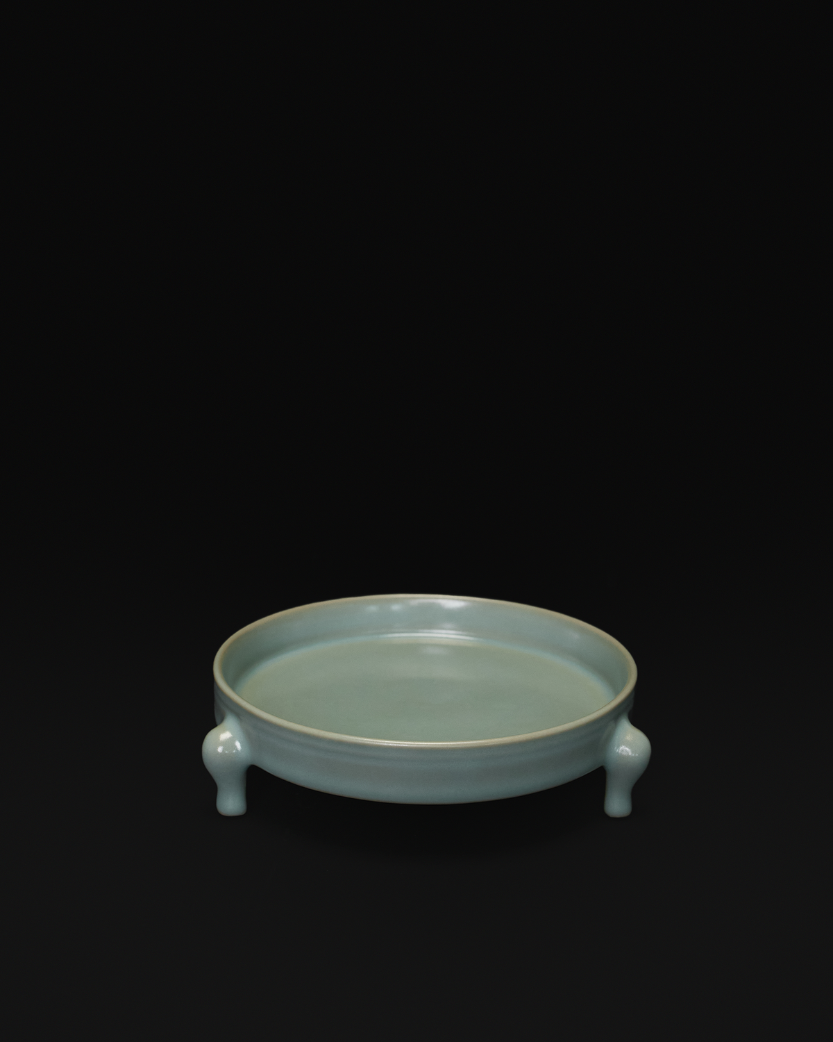 Three-quarter view of the celadon tripod bowl showing its shallow interior and three feet.