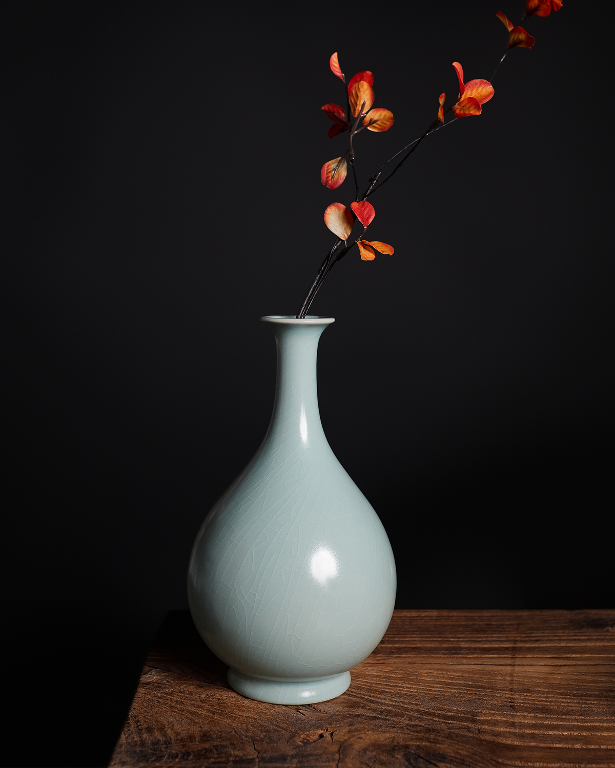 Celadon Yuhuchun vase holding red leafy branches on a wooden table against a dark background.