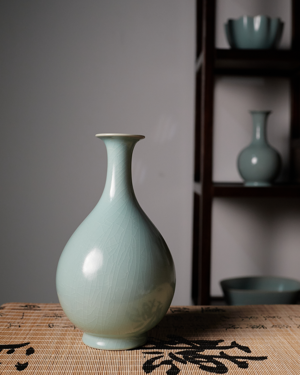 Yuhuchun vase displayed on a calligraphy-patterned surface with wooden shelving behind it.